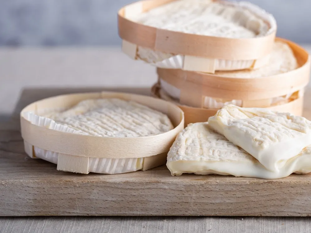 Pérail de Brebis (Pérail) cheese discs and wedges are stacked, displaying their wrinkled pale rind and smooth interior, all set on a wooden surface.