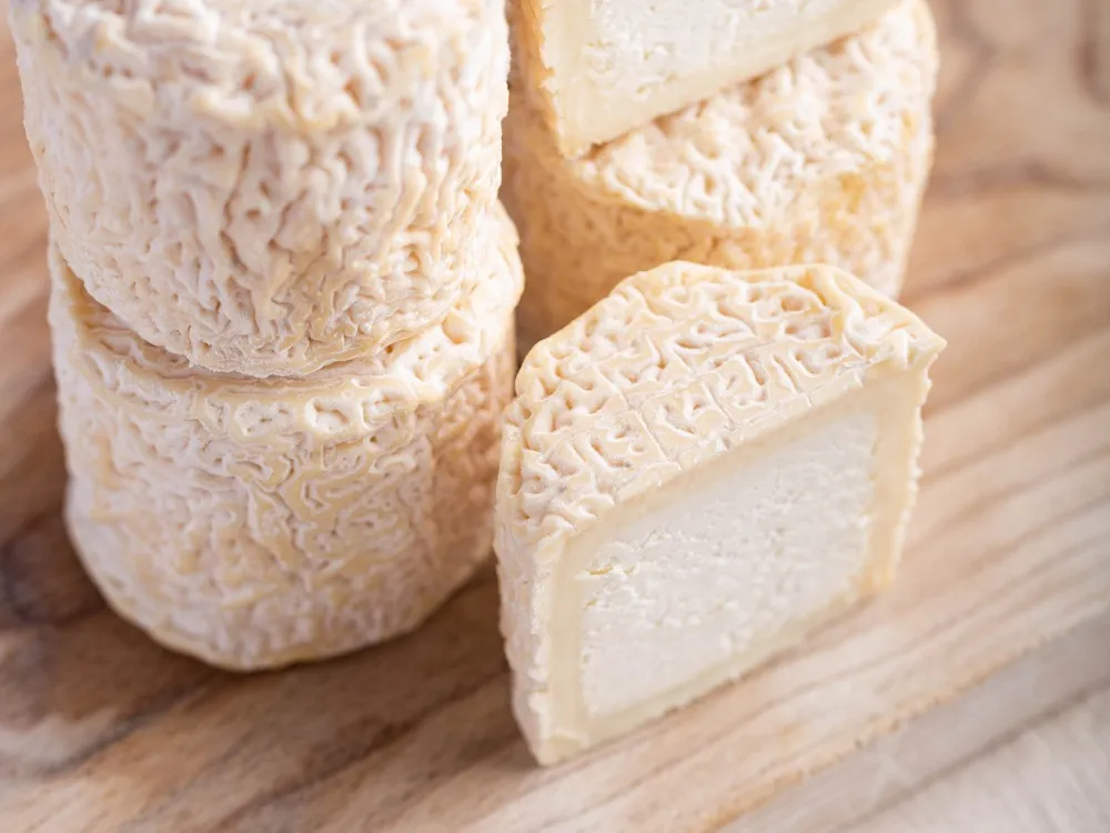 Small discs of Petit Blaja cheese, some stacked and others cut in half to reveal their creamy, soft texture and wrinkled yellow-orange rind, are arranged on a wooden surface