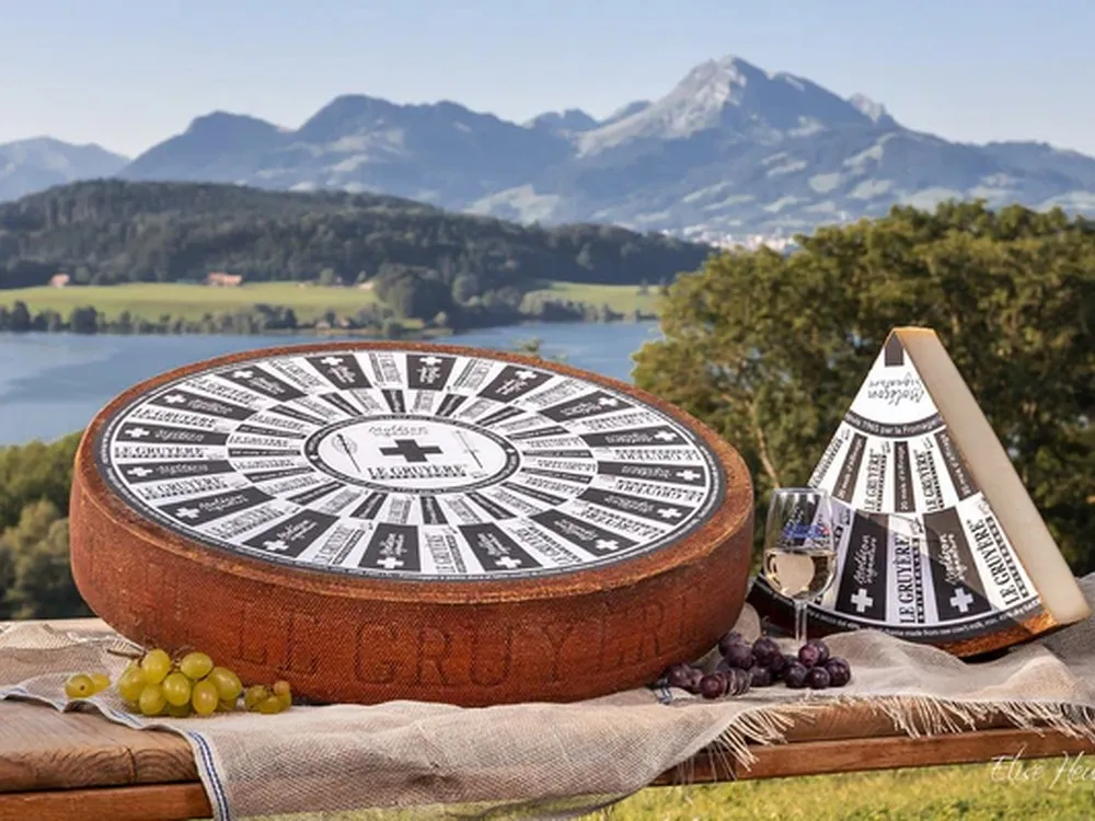 A wheel of Le Gruyère AOP cheese, accompanied by a triangular slice with a label on top, placed on a wooden board with a scenic backdrop of mountains and a river