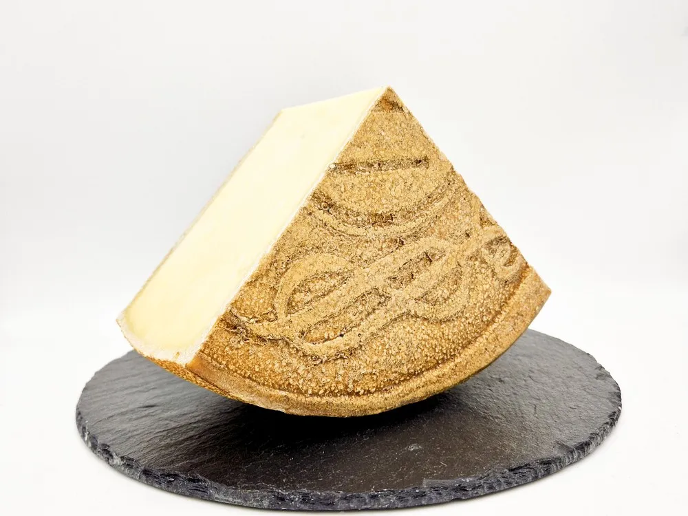 A slice of Alisia-Victoria cheese rests on a round marble surface, showcasing its golden-yellow rind and smooth interior