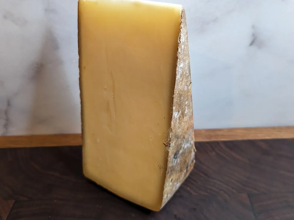 A wedge of Alpha Tolman cheese, showcasing its yellow interior and natural rind, rests on a wooden surface