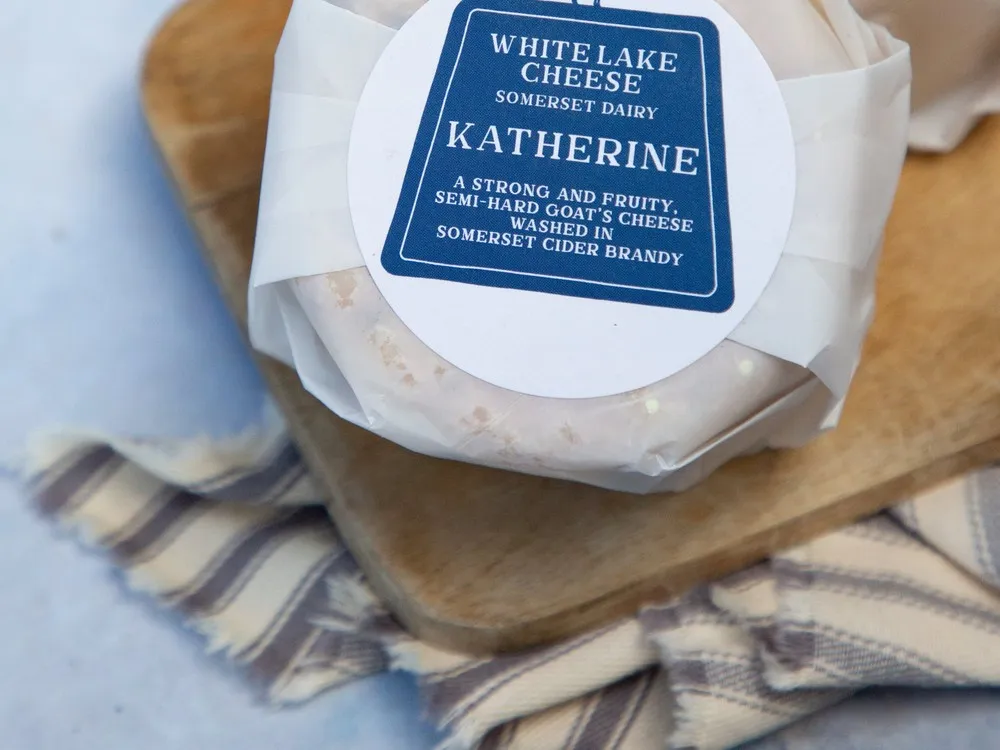 A disc of Baby Katherine cheese, wrapped in white paper with a label on top, rests on a wooden cutting board and a linen napkin