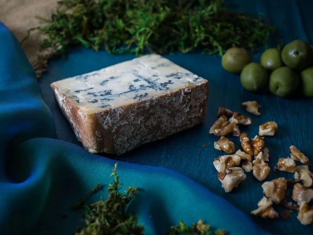 A wedge of Bayley Hazen Blue cheese, streaked with blue veins and encased in a dark brown rind, is elegantly arranged alongside fresh herbs, nuts, and olives, all resting on a rich, dark blue velvet cloth