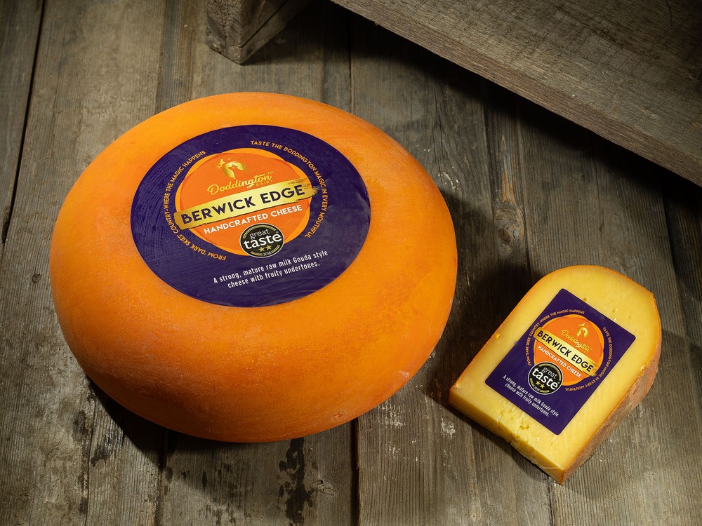 A wheel of Berwick Edge cheese, with its plastic rind, sits next to a slice of the cheese on a wooden surface