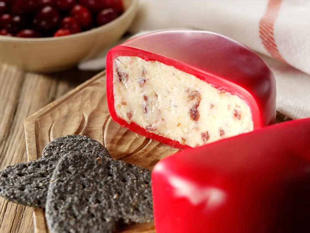 A half-cut small disc of Bouncing Berry cheese showcases its interior infused with flecks of cranberries, accompanied by crackers and fresh cranberries, all arranged on a wooden surface