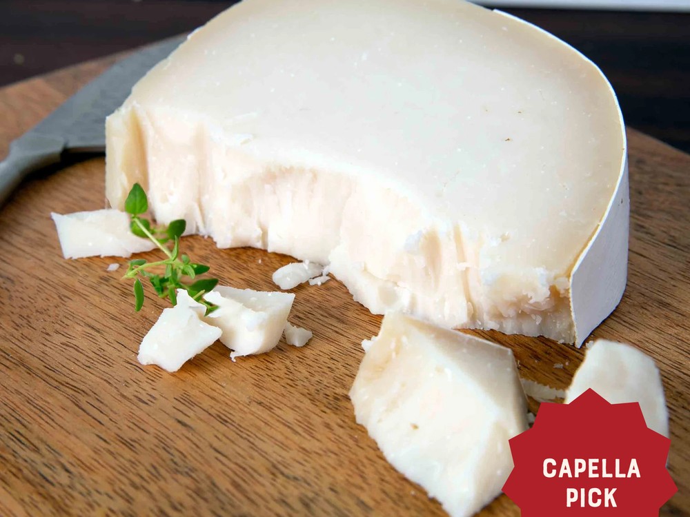 A partially sliced wheel of L'Amuse Brabander Goat Gouda, with chunks scattered on a wooden board, accompanied by fresh herbs and a cutting knife