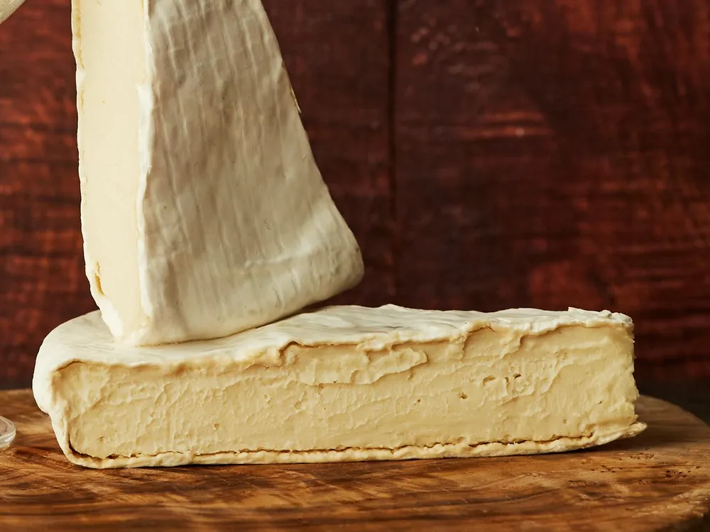 Triangular slices of Brick Lane Bree cheese stacked on a wooden surface, showcasing the white bloomy rind and soft, gooey interior.