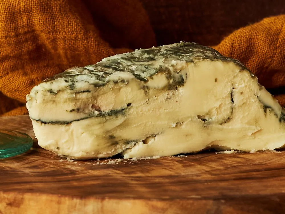 A piece of Brixton Blue, a vegan cheese, reveals blue veining on its yellow interior, highlighting its distinctive appearance and rich flavor