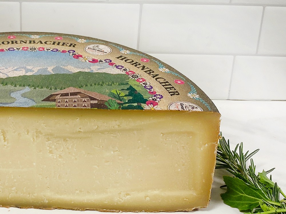 A partially cut wheel of Hornbacher cheese with its lable, rests against white tiled background, accompanied by fresh herbs