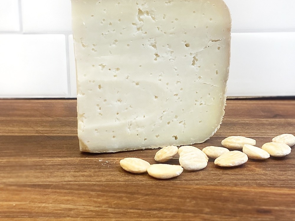 A slice of Ossau-Iraty cheese with an ivory-colored interior rests on a wooden surface, accompanied by blanched almonds