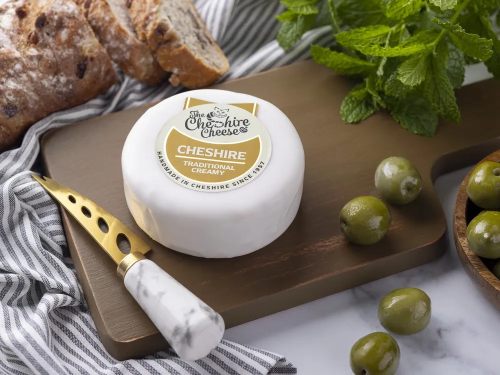 A creamy Cheshire cheese, wrapped in wax and labeled, rests on a wooden board with a knife, slices of bread, olives, and fresh herbs