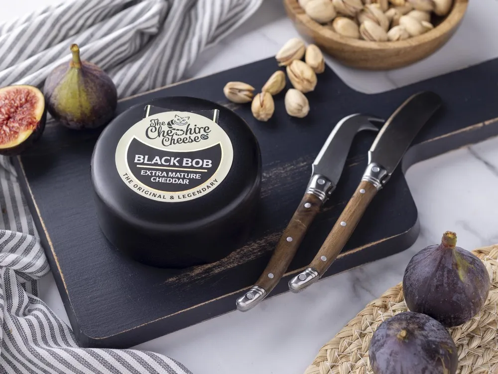 Black Bob Extra Mature Cheddar, encased in black wax and labeled, sits on a wooden cutting board alongside a knife, fresh figs, and nuts.