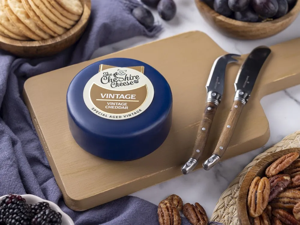 A wax-coated Vintage Special Aged Mature Cheddar with a label sits on a wooden board, surrounded by cheese knives, fresh fruits, nuts, berries, and cracker