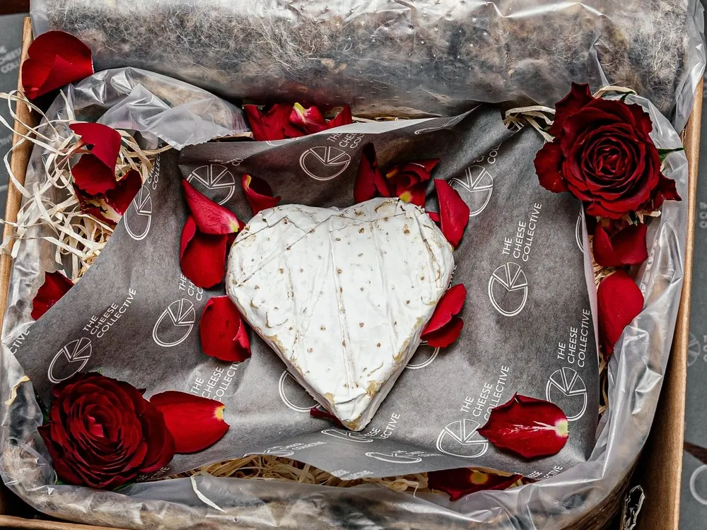 A heart-shaped Clava Brie, adorned with a white bloomy rind, rests in a basket surrounded by red roses and petals