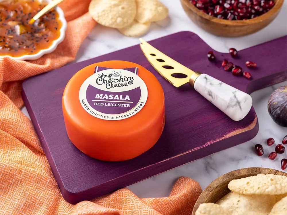 Masala - Spiced Mango Chutney & Nigella Seeds Red Leicester Cheese wrapped in red wax, placed on a purple wooden board with a knife, chutney, crackers, and pomegranate seeds