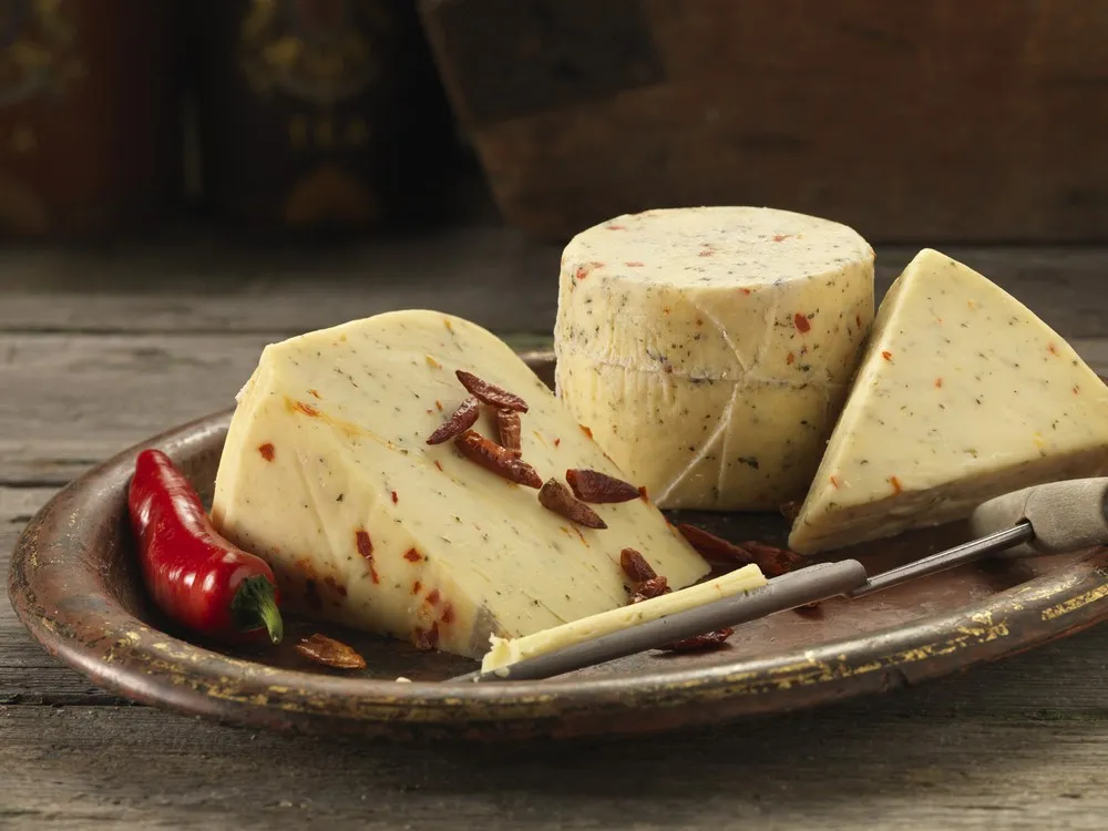 A cylindrical wheel of Northumberland Chilli cheese, with large slices revealing chili flecks, is arranged on a round wooden plate. It’s accompanied by a conical cheese sampler and red chilies, all set on a wooden surface