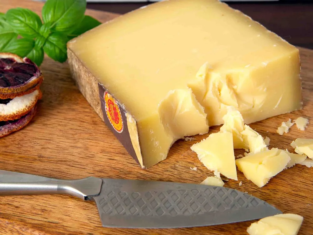 A slice of Comté cheese, along with several chunks, rests on a wooden board, accompanied by a cutting knife and a scattering of fresh herbs.