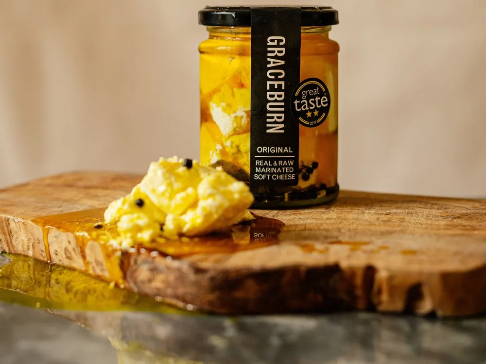 Graceburn cheese, dipped in oil with garlic and herbs and packed in a glass bottle, is accompanied by portions of the cheese and oil on a wooden surface