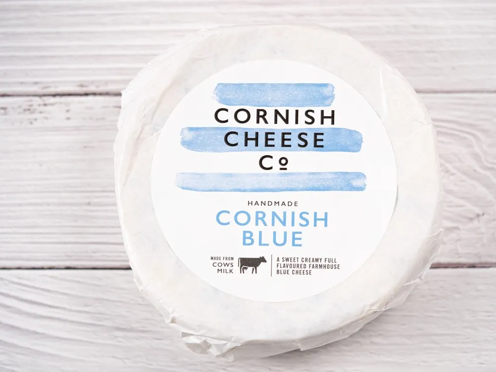 A Cornish Blue cheese wheel wrapped in white paper with a label, set against a white wooden board