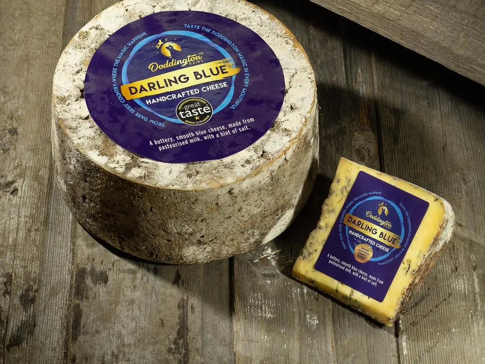 A wheel of Darling Blue cheese with a label sits next to a slice, showing its mold-ripened rind and blue-grey color, on a wooden surface