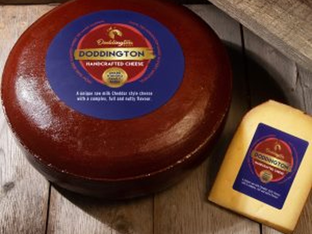 A wheel of Doddington cheese, with its plastic rind, sits next to a slice of the cheese on a wooden surface