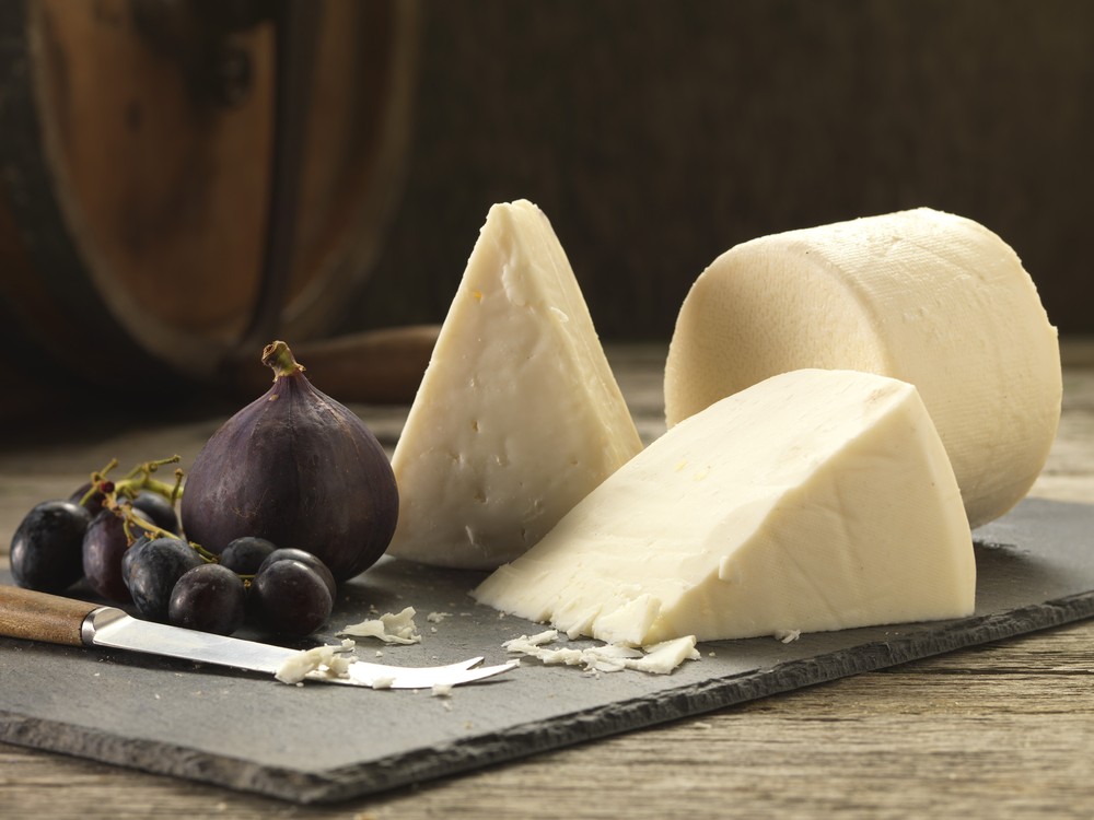 A cylindrical wheel of Elsdon cheese, with large slices, figs, black grapes, and a knife, sits on a black marble slab, all arranged on a wooden surface