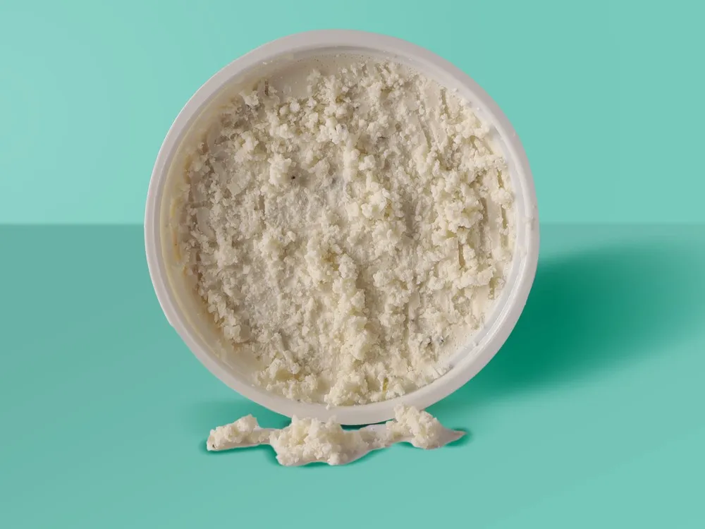 Garlic and Herb is a plant-based cheese in a plastic container, showcasing a creamy and crumbly texture