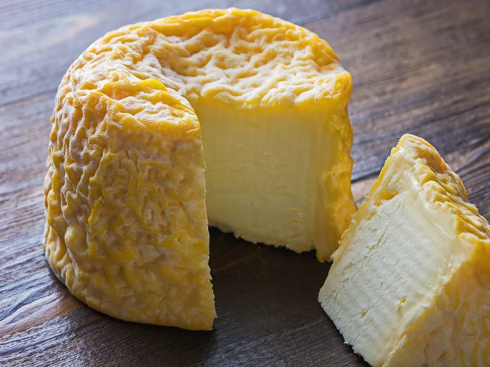 A cylindrical Langres cheese with a partially cut slice reveals its distinctive orangey-brown, wrinkled rind, resting on a wooden surface