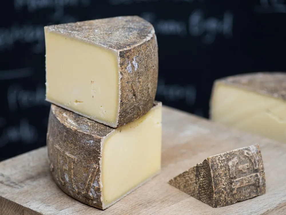 Triangular pieces of Le Maréchal cheese, stacked on a wooden surface, reveal a dark brown rind and a smooth, buttery interior