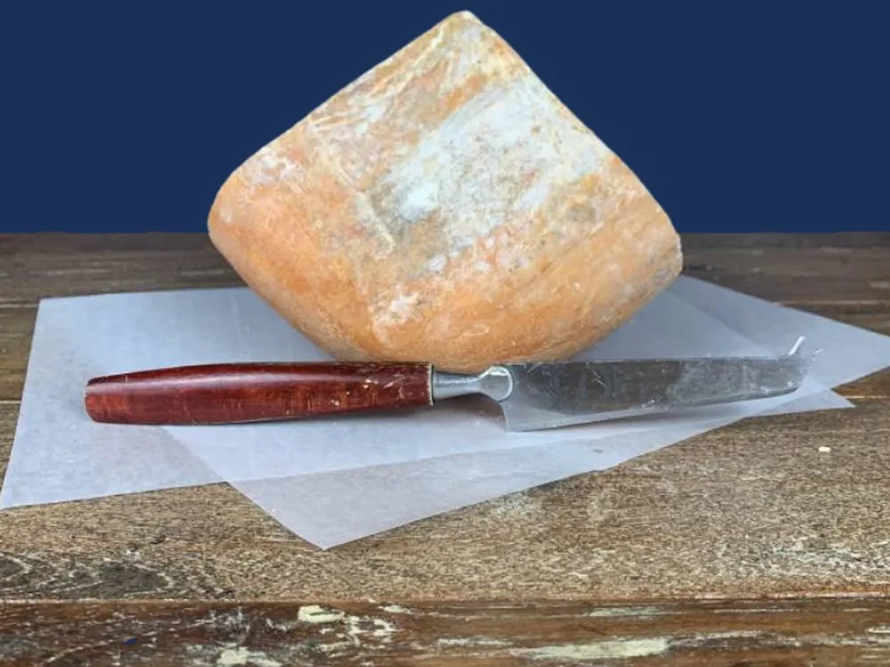 A piece of Mahón cheese, accompanied by a sharp knife, rests on butter paper placed on a wooden surface