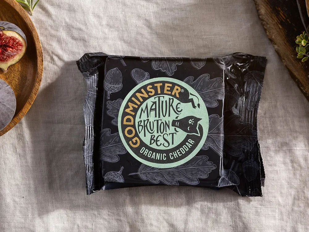 Godminster Mature Bruton Best Organic Cheddar, wrapped in its paper, rests on a white cloth alongside fresh figs