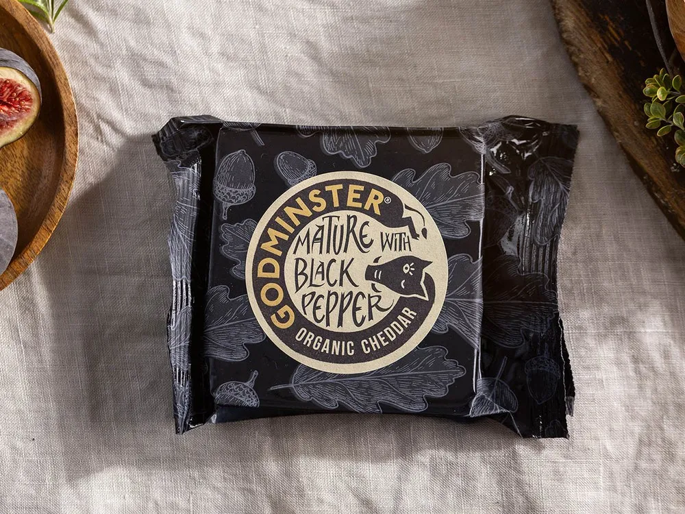 Godminster Mature Bruton Best with Black Pepper Organic Cheddar, wrapped in its paper, rests on a white cloth alongside fresh figs