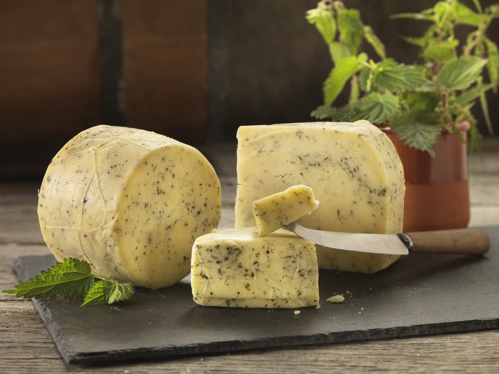 A cylindrical Northumberland Nettle cheese, along with its slices revealing a cream-colored interior speckled with herbs, is displayed on a black surface. Fresh nettle leaves and a knife on a wooden board accompany the arrangement