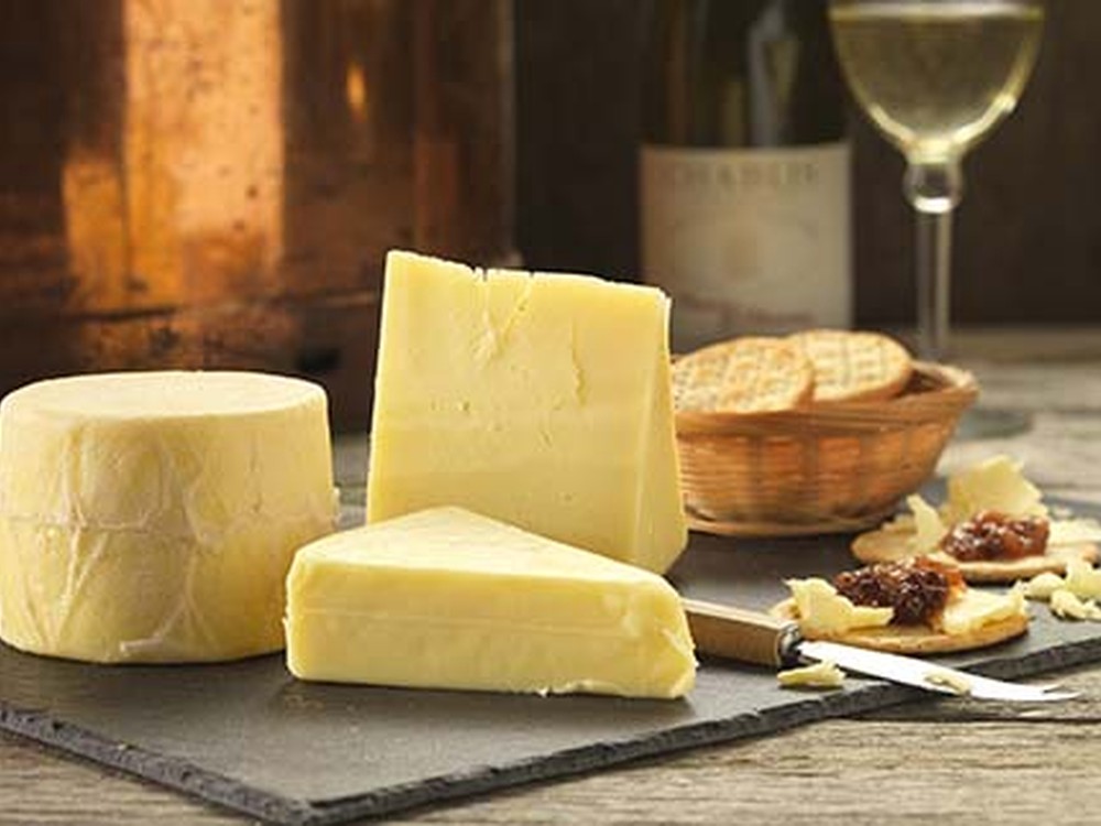 A cylindrical wheel of Northumberland Original Cheese, along with large slices, is arranged on black marble, accompanied by a knife, crackers, and a glass of white wine