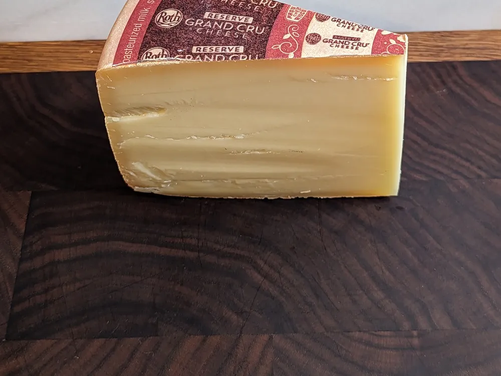 A wedge of wheel-shaped Grand Cru Reserve cheese, with its wrapper revealing a smooth interior, sits on a wooden surface