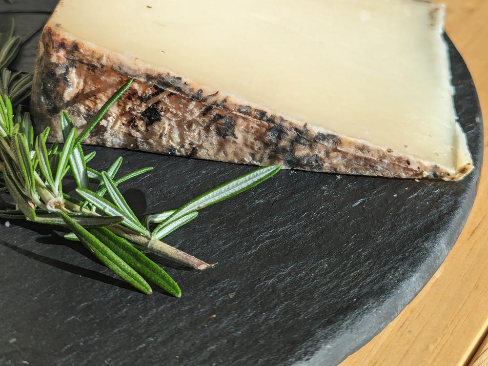 A wedge of Cave Aged Marisa cheese with a dark brown rind rests on a black marble surface, accompanied by fresh herbs.