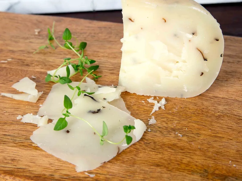 A slice and chunks of Truffle Pecorino (Pecorino al Tartufo) cheese, speckled with delicate truffle flecks, are displayed on a wooden surface, accompanied by fresh herbs