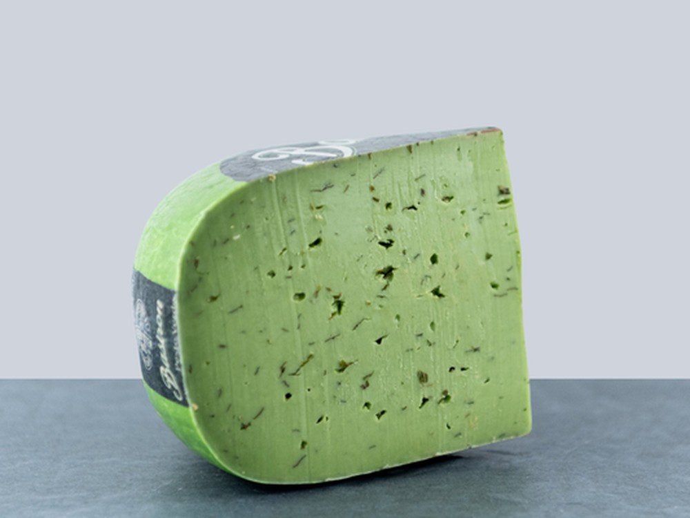 A wheel of Pesto Verde cheese with a slice cut out, showing green interior with basil and garlic flecks