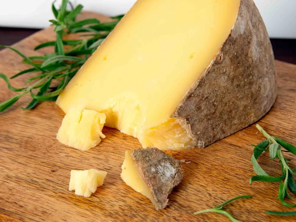 A slice of Pleasant Ridge Reserve cheese resting on a wooden board, surrounded by fresh herbs and small chunks of the cheese