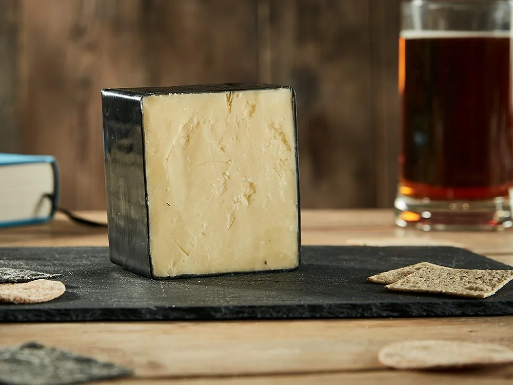 A cube of Pong's Extraordinary…Cheddar cheese, coated in wax, rests on a black surface alongside a book and crackers