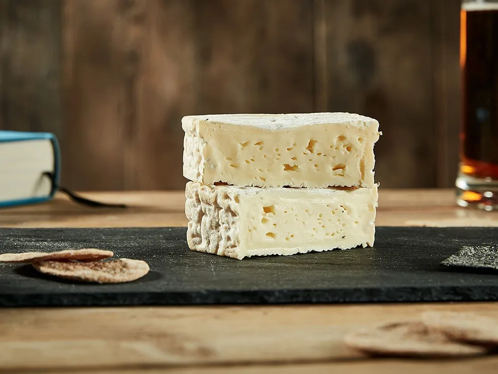 Rectangular pieces of Pont l'Eveque Cheese, stacked to reveal a pale yellow interior, sit on a wooden surface alongside crackers and a glass of beer