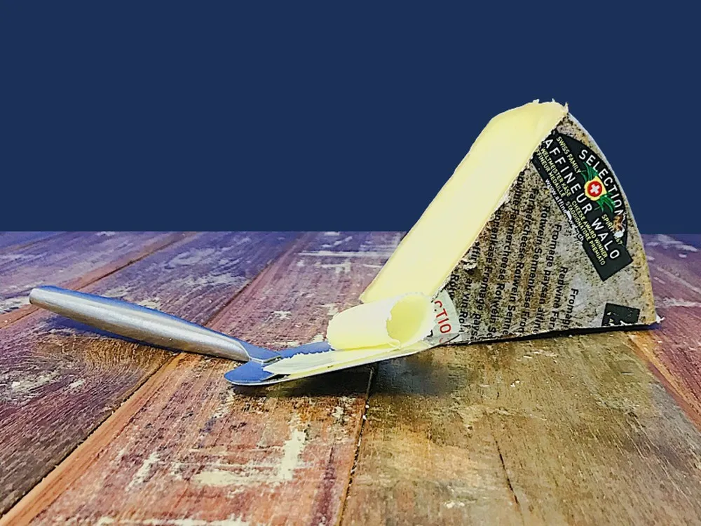 A slice of Affineur Walo Rotwein Sennechäs cheese, with a cheese slicer beside it, rests on a wooden surface