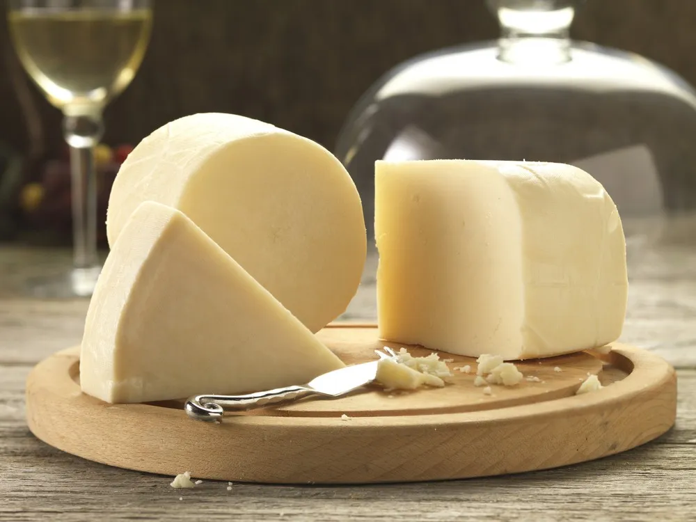 A cylindrical wheel of Redesdale cheese, accompanied by large slices and a knife, rests on a round wooden board, with a glass of white wine visible in the background