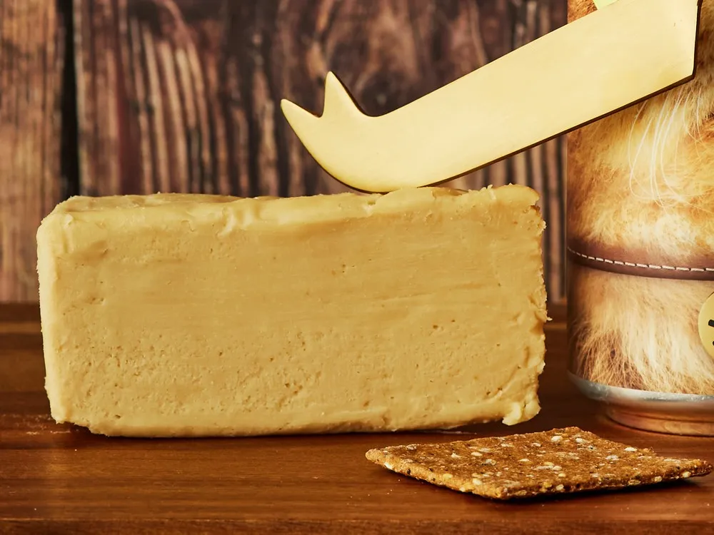 A block of Shoreditch Smoked cheese, showing its pale yellow color, alongside a cracker, rests on a wooden surface