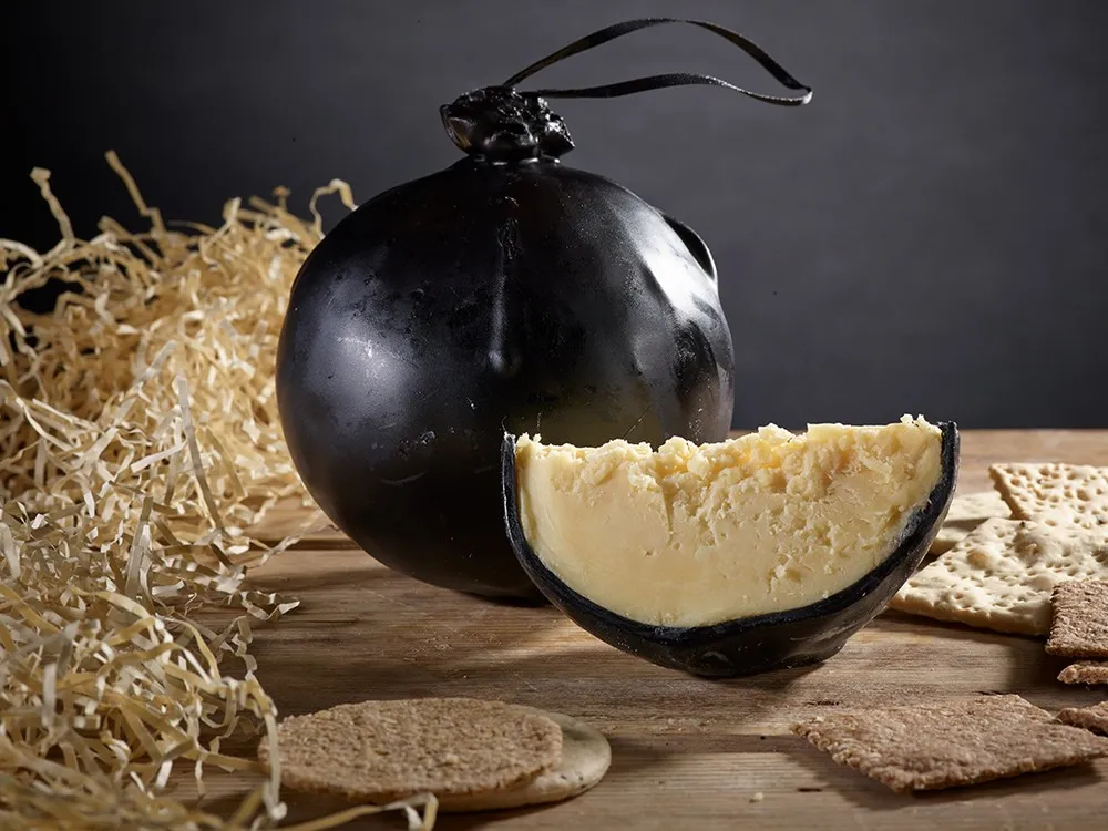 Shorrock's Lancashire Bomb cheese, encased in black wax, with a partially cut piece revealing its smooth interior, is accompanied by crackers and shredded paper, all set on a wooden surface