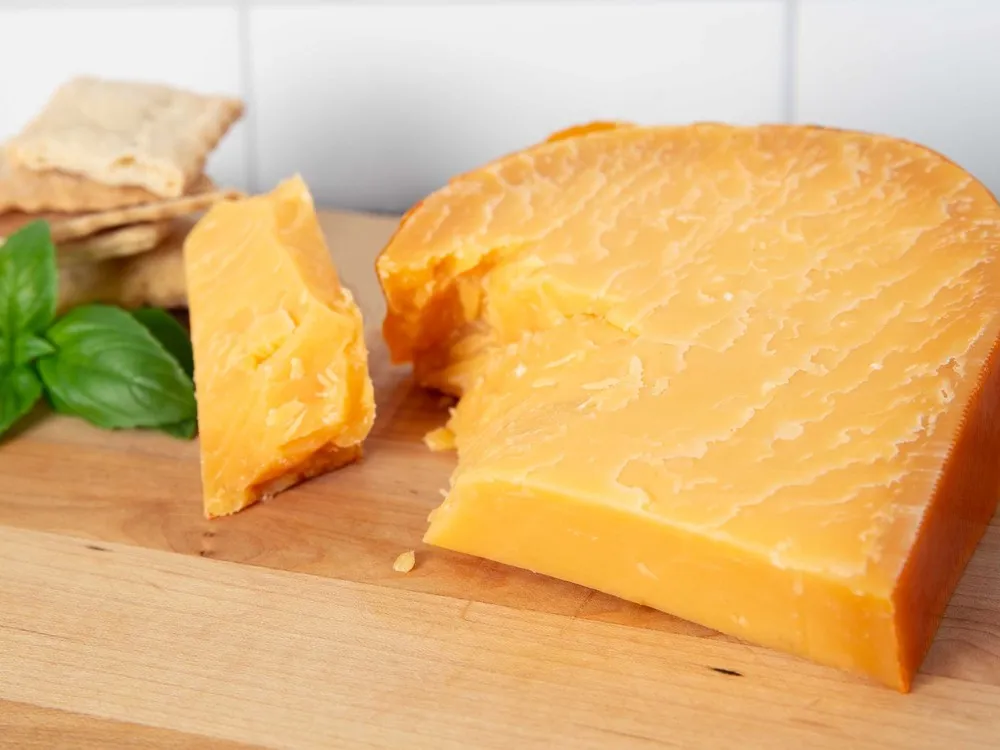 A slice of L'Amuse Signature Gouda displayed on a wooden surface, accompanied by fresh herbs and crackers