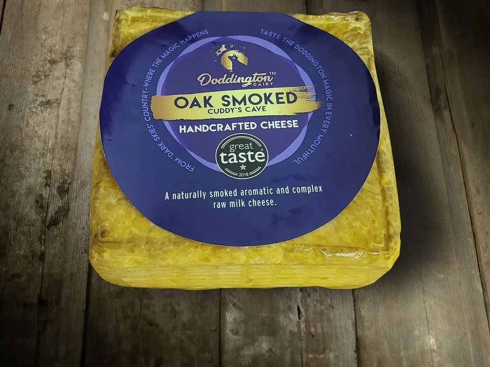 A rectangular block of Smoked Cuddy's Cave Cheese, wrapped in a plastic covering with a blue label, rests on a wooden surface