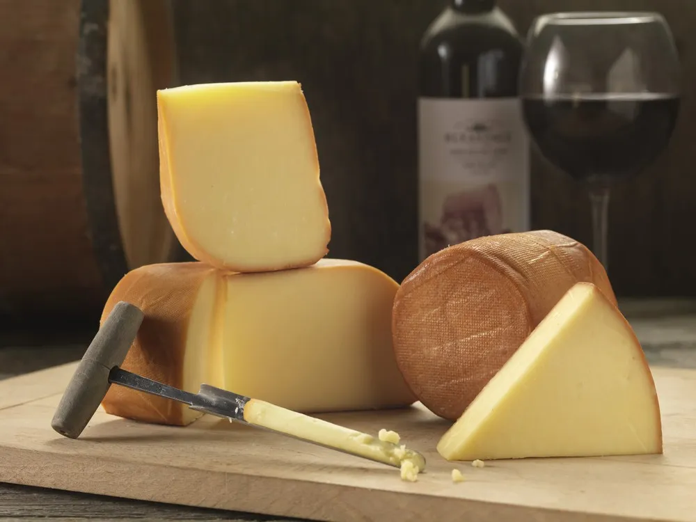 Slices of Northumberland Oak-smoked cheese in various sizes, showcasing their bronzed outer rind, are arranged on a wooden surface alongside a conical cheese sampler, a glass of red wine, and a wine bottle