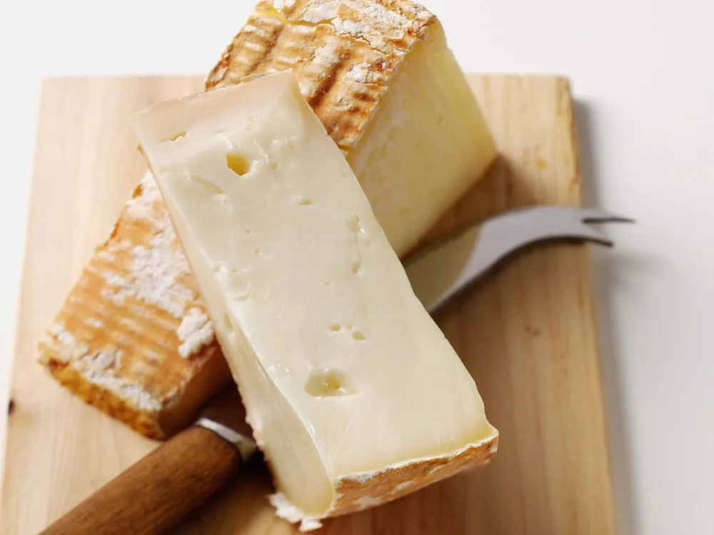 Wedges of Taleggio cheese with a creamy yellow texture and orange rind sits next to a cheese knife, showcasing its soft interior and wrinkled rind.