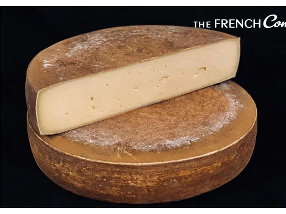 A half-cut wheel of Tomme du Jura cheese sits atop another, both showcasing their orange-colored rind and smooth interior, set against a black background